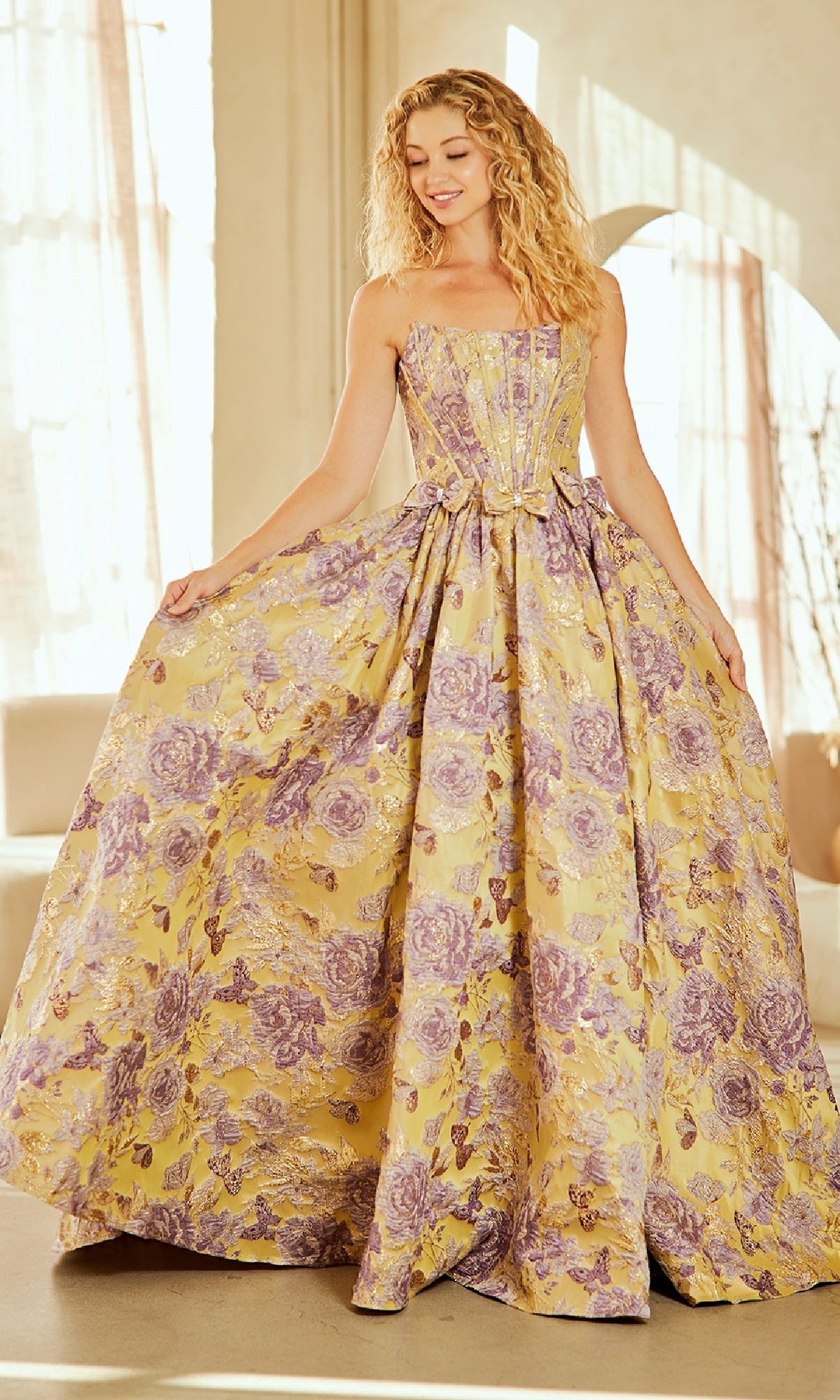 A woman in the Amelia Couture 8068 Lace-Up Print Prom Ball Gown, a strapless floor-length yellow dress with purple floral patterns, stands indoors gracefully holding her skirt's sides.